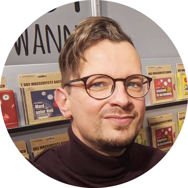 Mann mit braunen Haaren, leichtem Bart und Brille schaut in die Kamera und lächelt