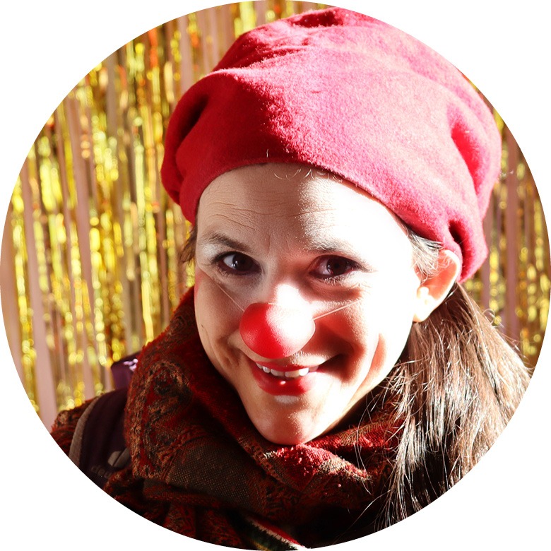 Frau mit roter Clownsnase und roter Mütze vor einem goldenen Vorhang